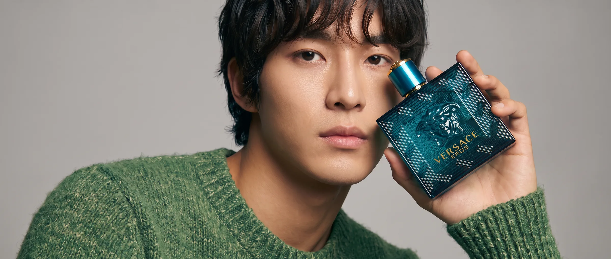 A man in a green knitted jumper holding a bottle of Versace Eros against his cheek