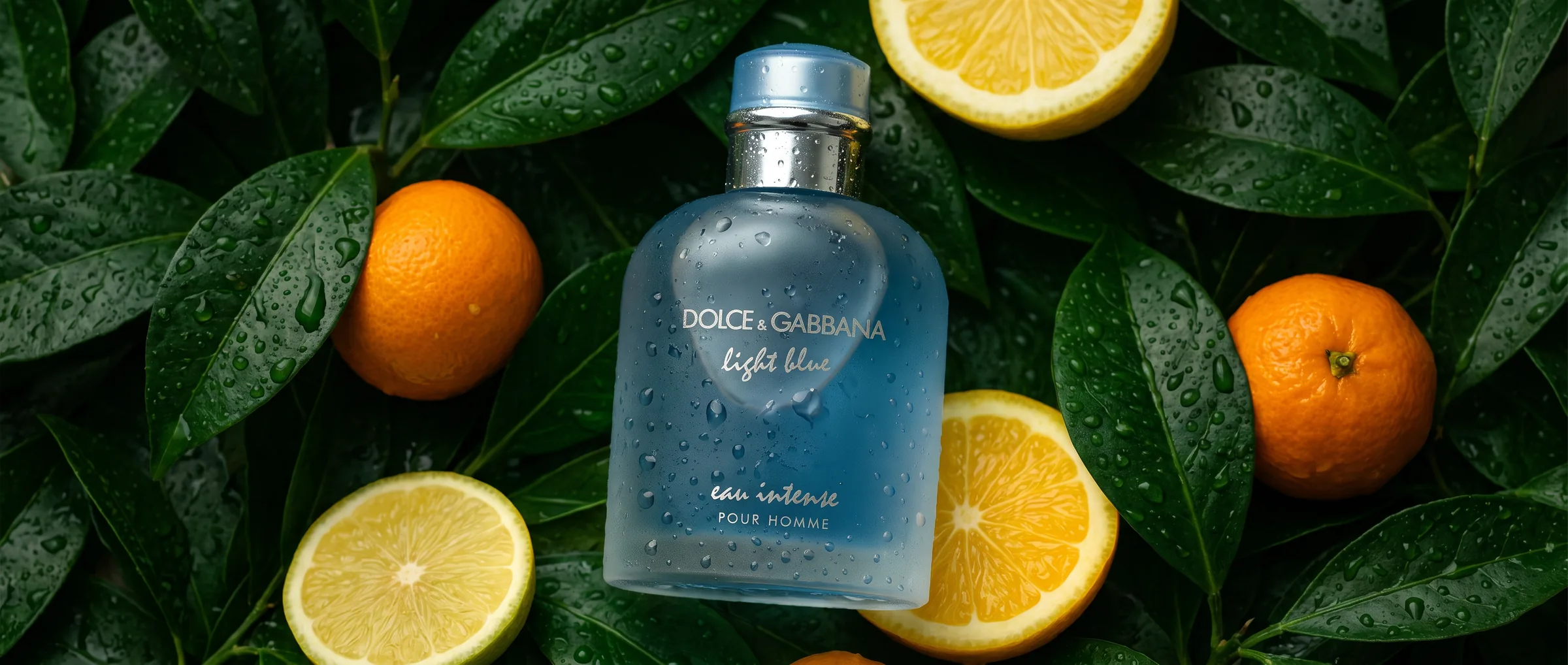 A frosted blue bottle of Dolce & Gabbana Light Blue Eau Intense Pour Homme lying among wet green leaves, lemons and oranges