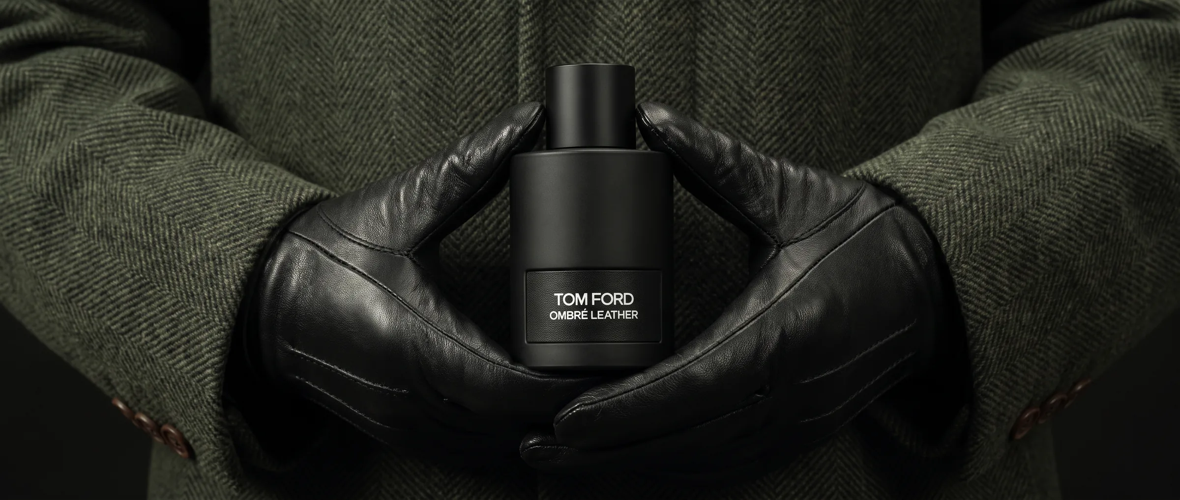 Black-gloved hands holding a bottle of Tom Ford Ombré Leather against a dark green wool coat