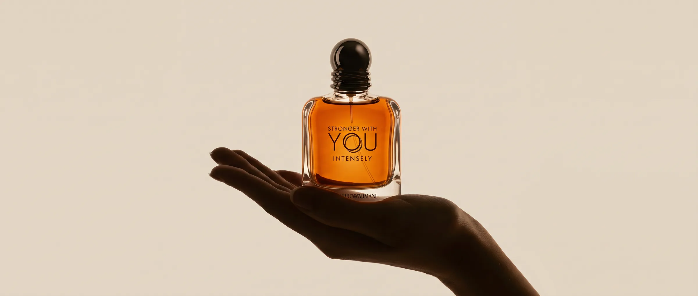 A hand holding up a bottle of Emporio Armani Stronger With You Intensely against a warm sand background