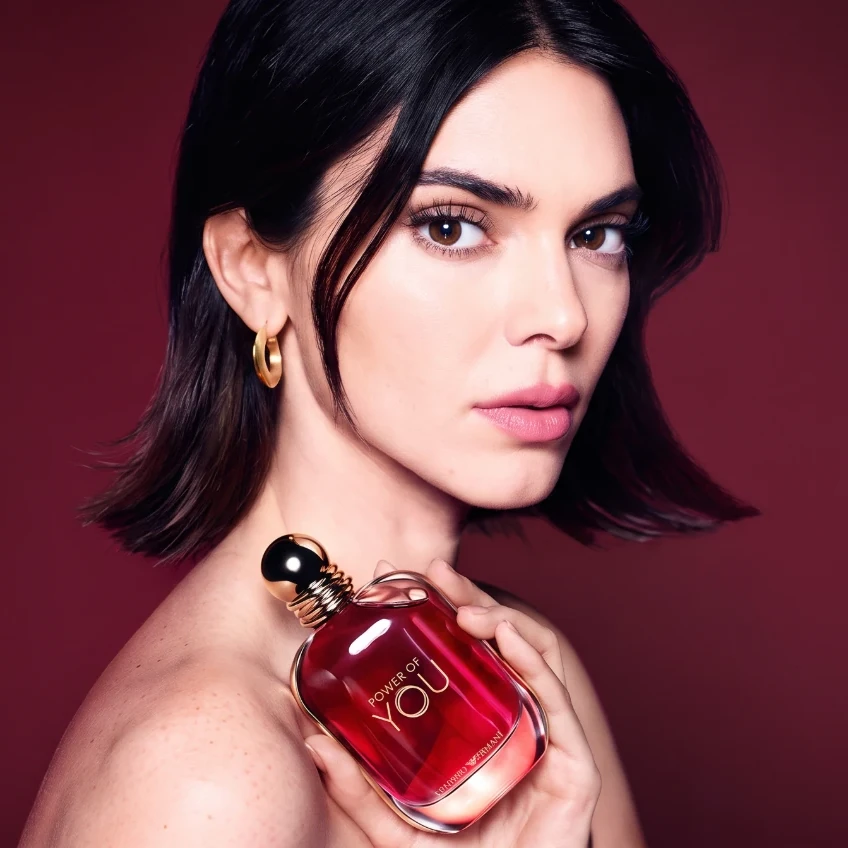 A woman with a dark bob holding a red bottle of Emporio Armani Power of You against her bare shoulder, on a deep red background